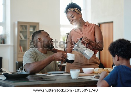 Young happy African female pouring coffee or tea into mug of her husband by table in front of their cute little son having breakfast Royalty-Free Stock Photo #1835891740