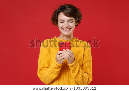 Smiling cheerful pretty attractive young brunette woman 20s wearing casual yellow sweater standing using mobile cell phone typing sms message isolated on bright red colour background studio portrait