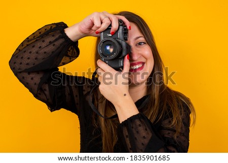 Portrait of fashionable young photographer woman over yellow background, taking pictures with vintage film camera.
