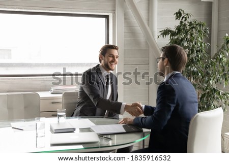 Happy young manager shaking hands with leader, feeling thankful for new job offer. Satisfied with good working results ceo executive manager praising male employee at meeting, sitting at table. Royalty-Free Stock Photo #1835916532