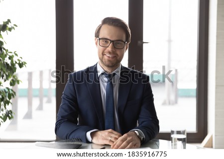 Portrait of happy young confident male leader in formal suit and glasses sitting at office table. Web camera view skilled ceo executive male manager holding online conversation negotiations meeting. Royalty-Free Stock Photo #1835916775