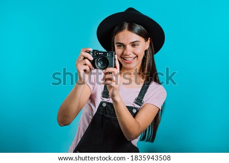 Young pretty woman in overall takes pictures with DSLR camera over blue background in studio. Girl smiling and having fun as photographer.