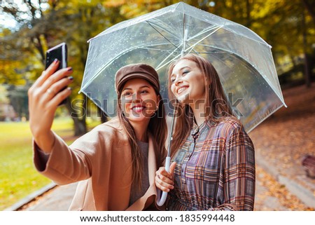 Outdoor portrait of two young women taking selfie using phone under umbrella during rain in autumn park.