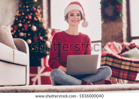 Photo of pretty young girl hold netbook sit carpet wear santa headwear red pullover jeans socks decorated living room indoors.
