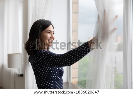 Happy young asian woman open curtains look in window distance dreaming or planning future success. Smiling Vietnamese female renter tenant meet welcome new sunny day in own house or apartment. Royalty-Free Stock Photo #1836020647