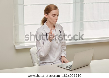 A young female doctor, in a medical gown, looks at the laptop screen and talks. Doctor's consultation online.
