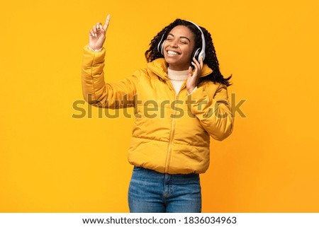 African Woman Listening And Enjoying Music In Wireless Earphones Standing Over Yellow Studio Background. Autumn Playlist, Winter Songs And Online Music Application Concept