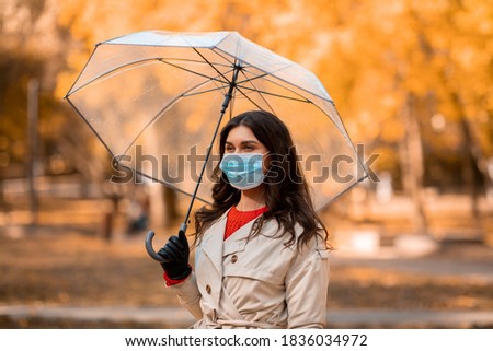 Young woman in protective mask walking with umbrella at park on autumn day. Millennial lady on walk under rain with face protection against coronavirus. Covid-19 fall concept