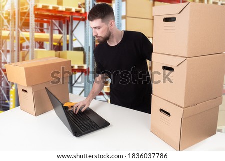 Storekeeper in the warehouse. Warehouse account. This man gives orders from the warehouse. The storekeeper works on a laptop. A man next to cardboard boxes and a laptop. Royalty-Free Stock Photo #1836037786