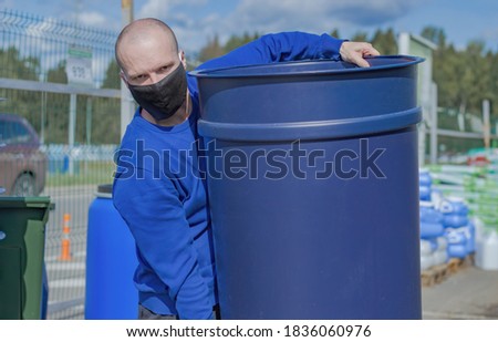 A Male wearing a medical mask carries a blue plastic container. In the background you can see pallets with building materials and an iron fence.