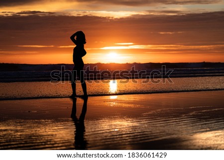Silhouette shadow shape of a girl is posting action on the beach with dramatic burning orange sky sunset environment. Relaxation and recreation concept photo.