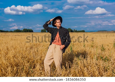 Rural Countryside Scene. Tall handsome man dressed in a black shirt and black hat standing at golden oat field. Summer landscape with blue sky