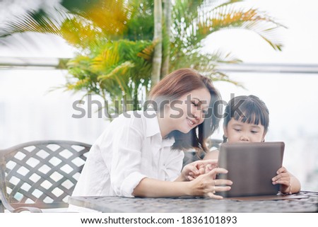 Young mother spending time with daughter in cafe and helping her to upload new application on digital tablet