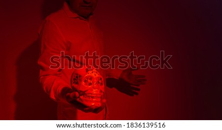 A man whose face is unrecognizable with a painted skull from the day of the dead or halloween in his hand. Photograph in red tones. Selective focus