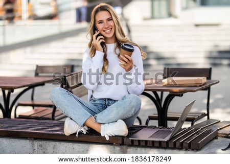 Cheerful young woman in casual clothes with legs crossed sitting on bench and talking on mobile phone while looking away in park