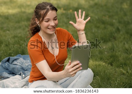 Young pretty smiling girl using headphones and tablet pc to speak with friends through video chat. Dark haired girl sitting on the green grass and use technologies for searching information.