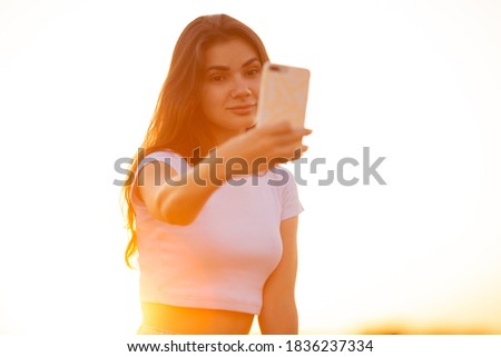 Young  caucasian woman in a whit t shirt making selfie sitting on stone at sunset. Happy girl writes a message or communicates on the Internet at sunny day. Lifestyle concept with copy space