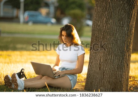 Young female, freelance woman in white t shirt working on laptop pc computer,  in city park on green grass sunshine lawn outdoors