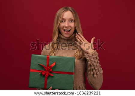 Image of charming young blonde womanin trendy knitted sweater smiling and holding gift with red ribbon. Studio shot red background. New Year Women's Day birthday holiday concept