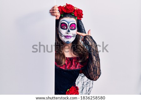Young woman wearing day of the dead custome holding blank empty banner pointing with hand finger to face and nose, smiling cheerful. beauty concept 