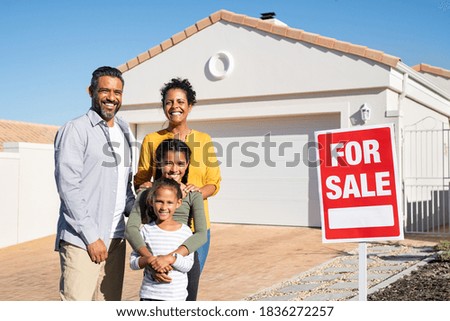 Happy mixed race family standing outside home with sale signboard. Couple with two daughters standing by for sale sign outside their new house while looking at camera, put the house up for sale.