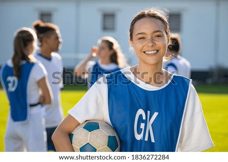 Cheerful soccer player holding a football and looking at camera. Portrait of young woman during training on soccer field. Satisfied high school student with her teammates standing in background. Royalty-Free Stock Photo #1836272284