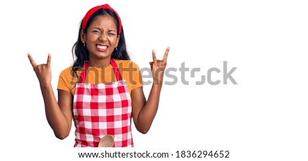 Young indian girl wearing professional baker apron shouting with crazy expression doing rock symbol with hands up. music star. heavy concept. 