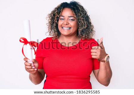 Young african american plus size woman holding graduate degree diploma smiling happy and positive, thumb up doing excellent and approval sign 