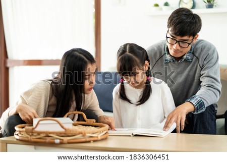 Asian cute girl reading story cartoon book with her mom and dad in living room. Happiness cheerful family and domestic lifestyle concept.