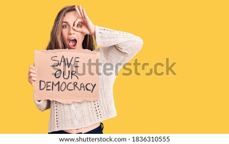 Young beautiful blonde woman holding save our democracy banner smiling happy doing ok sign with hand on eye looking through fingers 