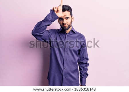 Young handsome man with beard wearing casual shirt making fun of people with fingers on forehead doing loser gesture mocking and insulting. 