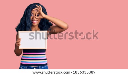 Beautiful african american woman holding blank empty banner smiling happy doing ok sign with hand on eye looking through fingers 