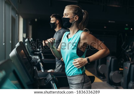 Young fit woman and man running on treadmill in modern fitness gym. They keeping distance and wearing protective face masks. Coronavirus world pandemic and sport theme. Royalty-Free Stock Photo #1836336289