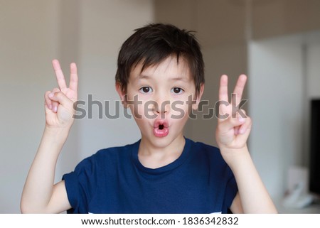 Cute boy making two finger and funny face at home. Mixed race Asian German kid headshot portrait victory sign. School child about 6-7 year old.