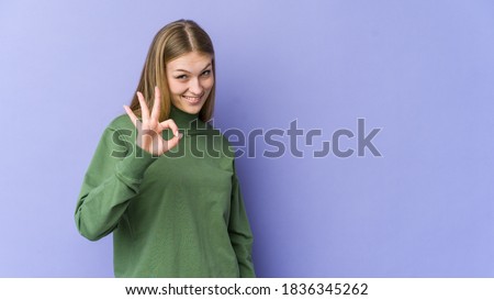 Young blonde woman isolated on purple background winks an eye and holds an okay gesture with hand.