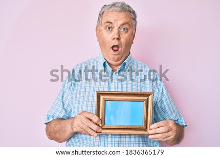 Senior grey-haired man holding empty frame scared and amazed with open mouth for surprise, disbelief face 