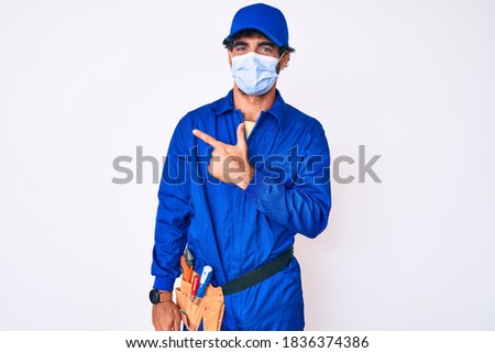 Handsome young man with curly hair and bear wearing handyman uniform and covid-19 safety mask smiling cheerful pointing with hand and finger up to the side 