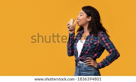 Time For Snack. Side View Portrait Of Happy Black Lady Eating Hamburger. African American Woman Taking Bite Of Tasty Fast Food, Isolated Over Orange Studio Background, Banner, Copy Space