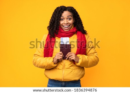 Joyful African Tourist Lady Holding Tickets And Passport Going On Winter Vacation Posing Over Yellow Studio Background, Smiling To Camera. Traveling On Christmas Holidays, Tourism Concept.