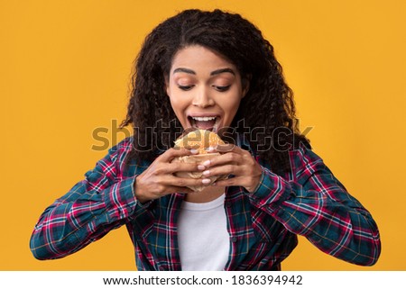 Junk Food. Closeup Portrait Of Funny Hungry African American Woman Eating Burger. Black Lady With Curly Hair Holding And Taking Bite Of Tasty Sandwich, Isolated Over Orange Studio Background, Banner