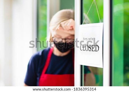 Owner hanging closed sign on shop entrance. Coronavirus outbreak and lockdown. Sad woman wearing face mask holding notice board next to restaurant or cafe door. Pandemic business recession.