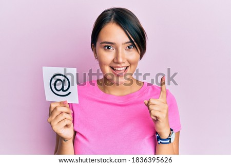 Beautiful young woman with short hair holding paper with email symbol smiling with an idea or question pointing finger with happy face, number one 