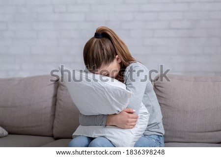 Young woman with depression hugging pillow and crying on sofa at home. Lonely millennial lady feeling stressed and hopeless, suffering from mood disorder or having psychological problem Royalty-Free Stock Photo #1836398248