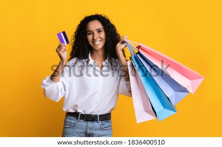 Happy brunette woman with colorful shopping bags and credit card standing on yellow background in studio, cheerful shopaholic lady enjoying seasonal sales and easy payments, got cashback, copy space