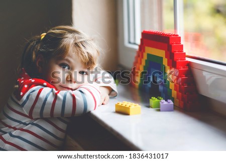 Cute little toddler girl by window create rainbow with colorful plastic blocks during pandemic coronavirus quarantine. Children made and paint rainbows around the world as sign.