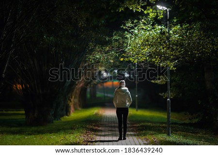 Young alone woman in white jacket walking on sidewalk through alley of trees under lamp light in autumn night. Spending time alone in nature. Peaceful atmosphere. Back view. Royalty-Free Stock Photo #1836439240