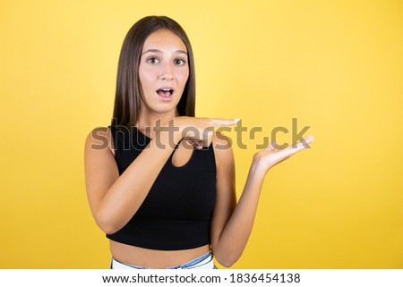 Beautiful young girl kid over isolated yellow background surprised, showing and pointing something that is on her hand