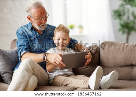 Happy family senior grandfather and boy sitting on sofa and watching cartoon on tablet on weekend day at home together
