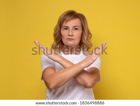 Mature woman making X sign with crossed hands, gesturing stop, warning of danger. No way, stop doing. Studio shot on yellow background