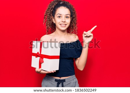 Beautiful kid girl with curly hair holding gift smiling happy pointing with hand and finger to the side 